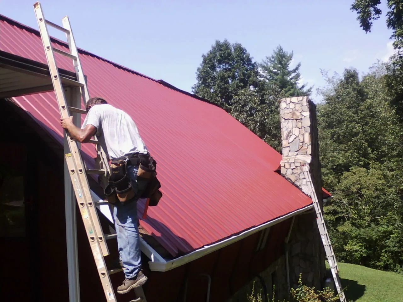 Roofer on a ladder beside a red metal roof and stone chimney — Soffit & Fascia Repair by Roofing Brothers in Norco
